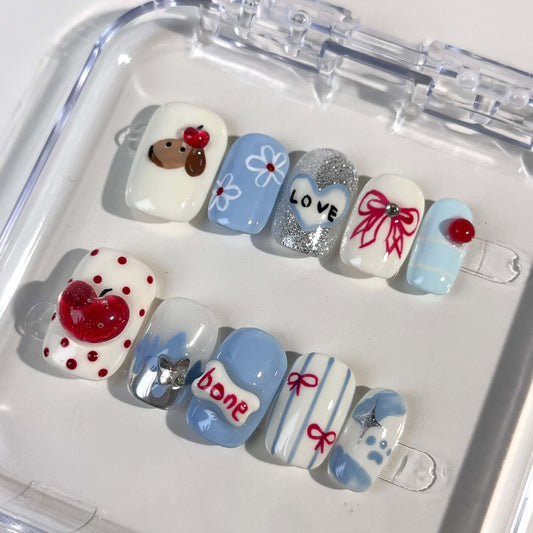 A set of press - on nails with a variety of cute and whimsical designs, including a dog, apple, heart with the word "LOVE", bow, bone, and sparkling elements, neatly arranged in a transparent case. The nails have a playful and charming finish, ideal for casual and fun - filled events.
