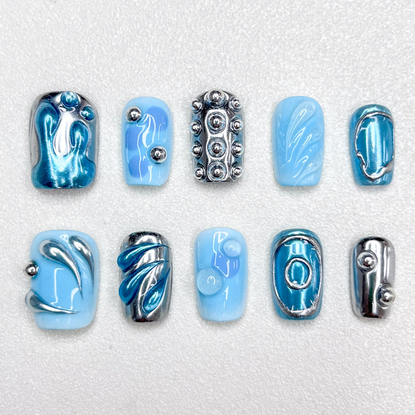 A cyber-inspired press-on nail set in icy blue and metallic chrome tones, featuring 3D orbs, abstract swirls, and smooth reflective patterns. A sleek, sci-fi style manicure with avant-garde vibes.