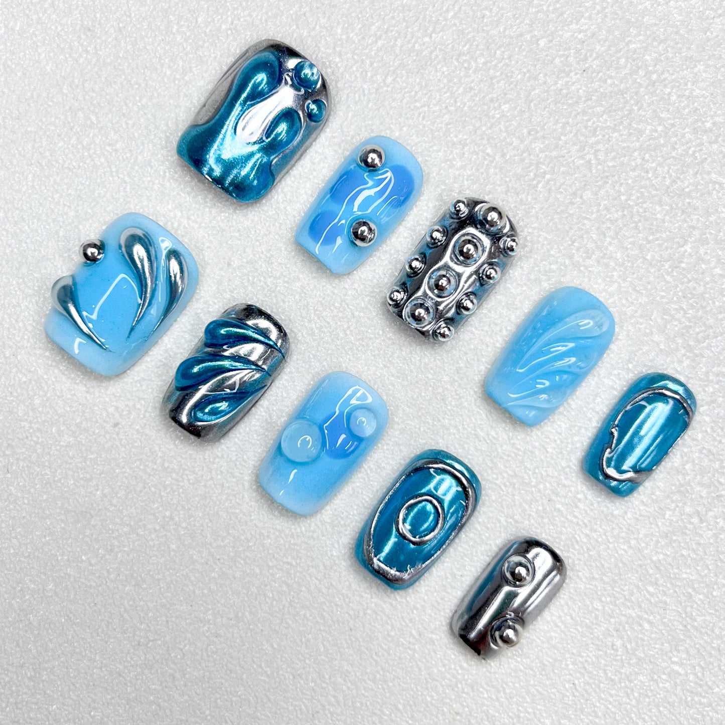 A cyber-inspired press-on nail set in icy blue and metallic chrome tones, featuring 3D orbs, abstract swirls, and smooth reflective patterns. A sleek, sci-fi style manicure with avant-garde vibes.