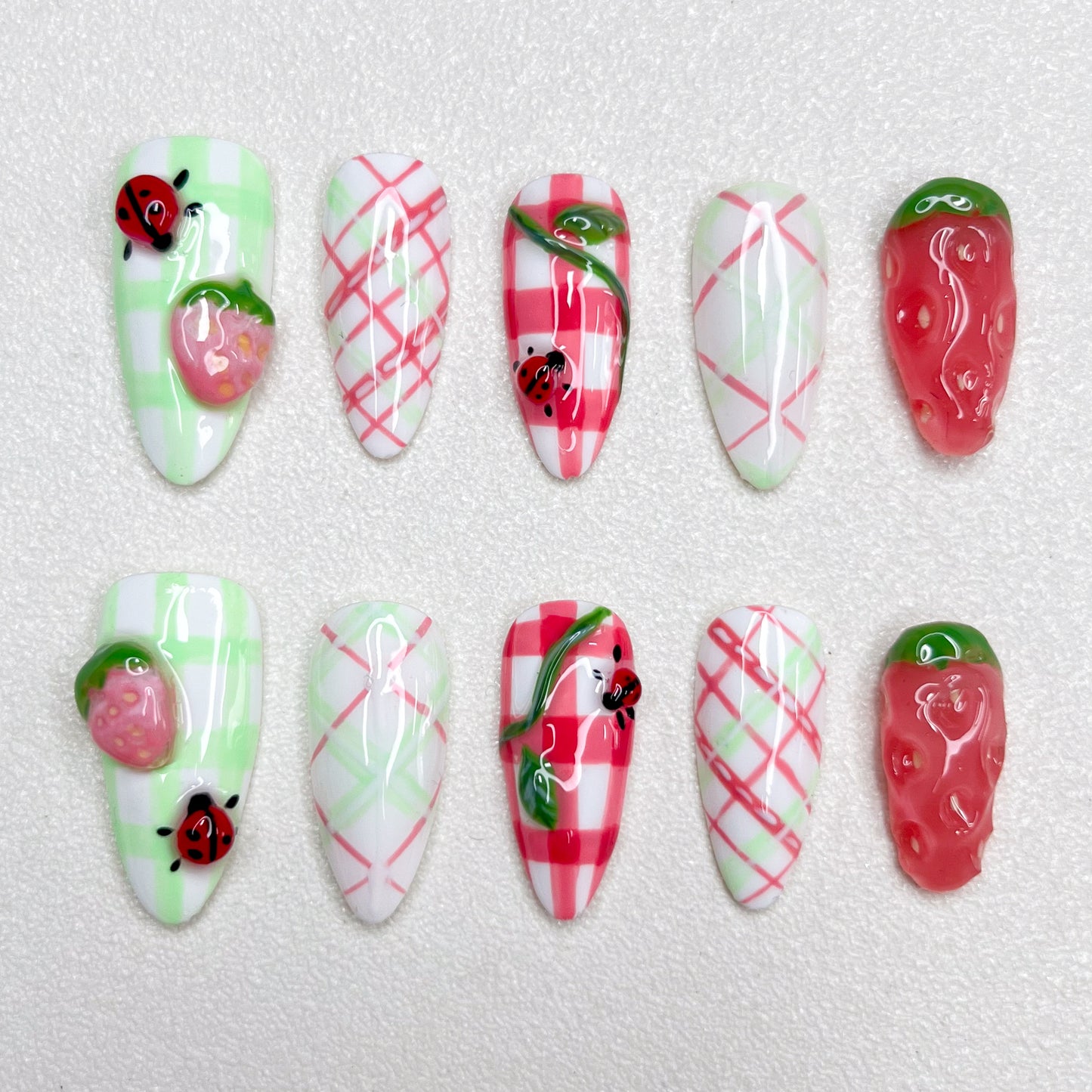 A charming press-on nail set decorated with checkered gingham prints, pink strawberries, and little ladybugs, combining fruity textures and playful summer picnic vibes in green, red, and white hues.