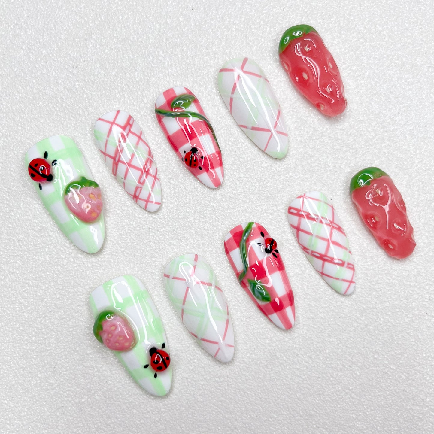 A charming press-on nail set decorated with checkered gingham prints, pink strawberries, and little ladybugs, combining fruity textures and playful summer picnic vibes in green, red, and white hues.