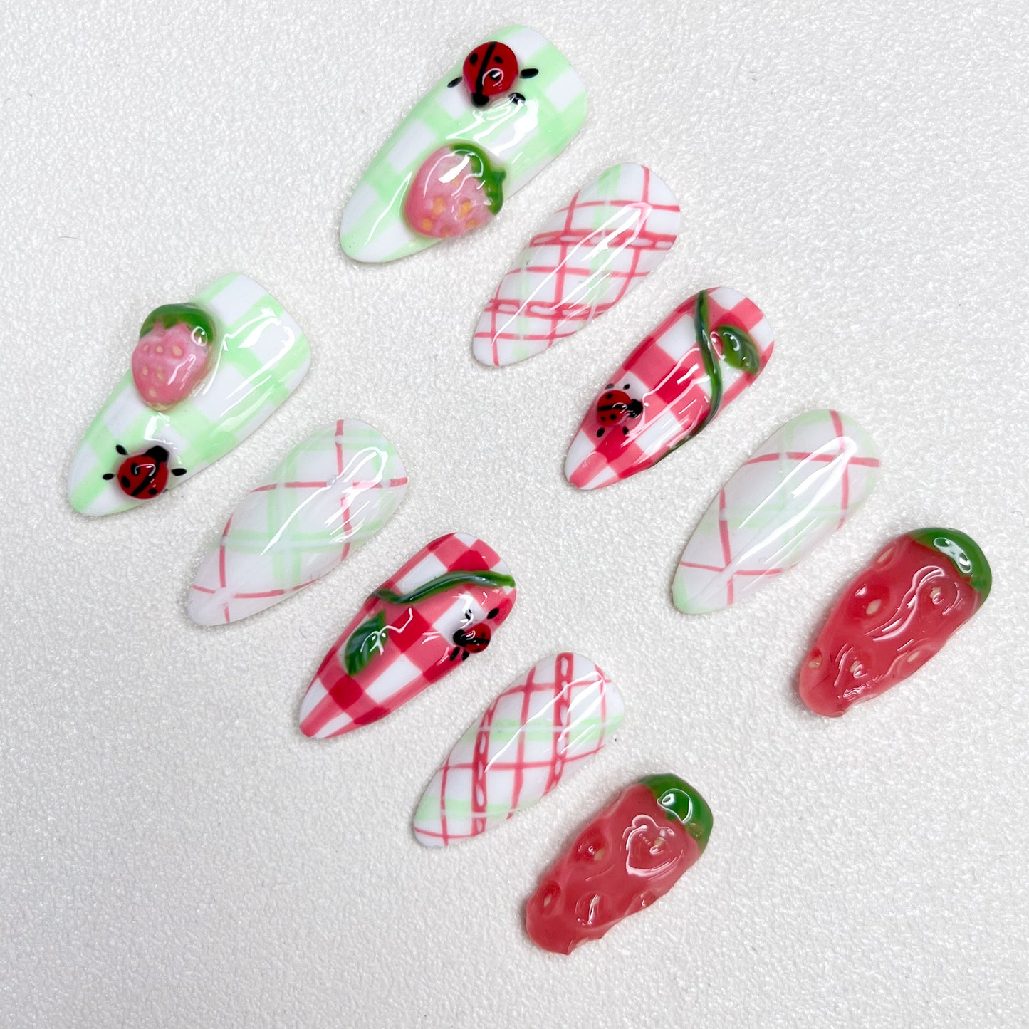 A charming press-on nail set decorated with checkered gingham prints, pink strawberries, and little ladybugs, combining fruity textures and playful summer picnic vibes in green, red, and white hues.
