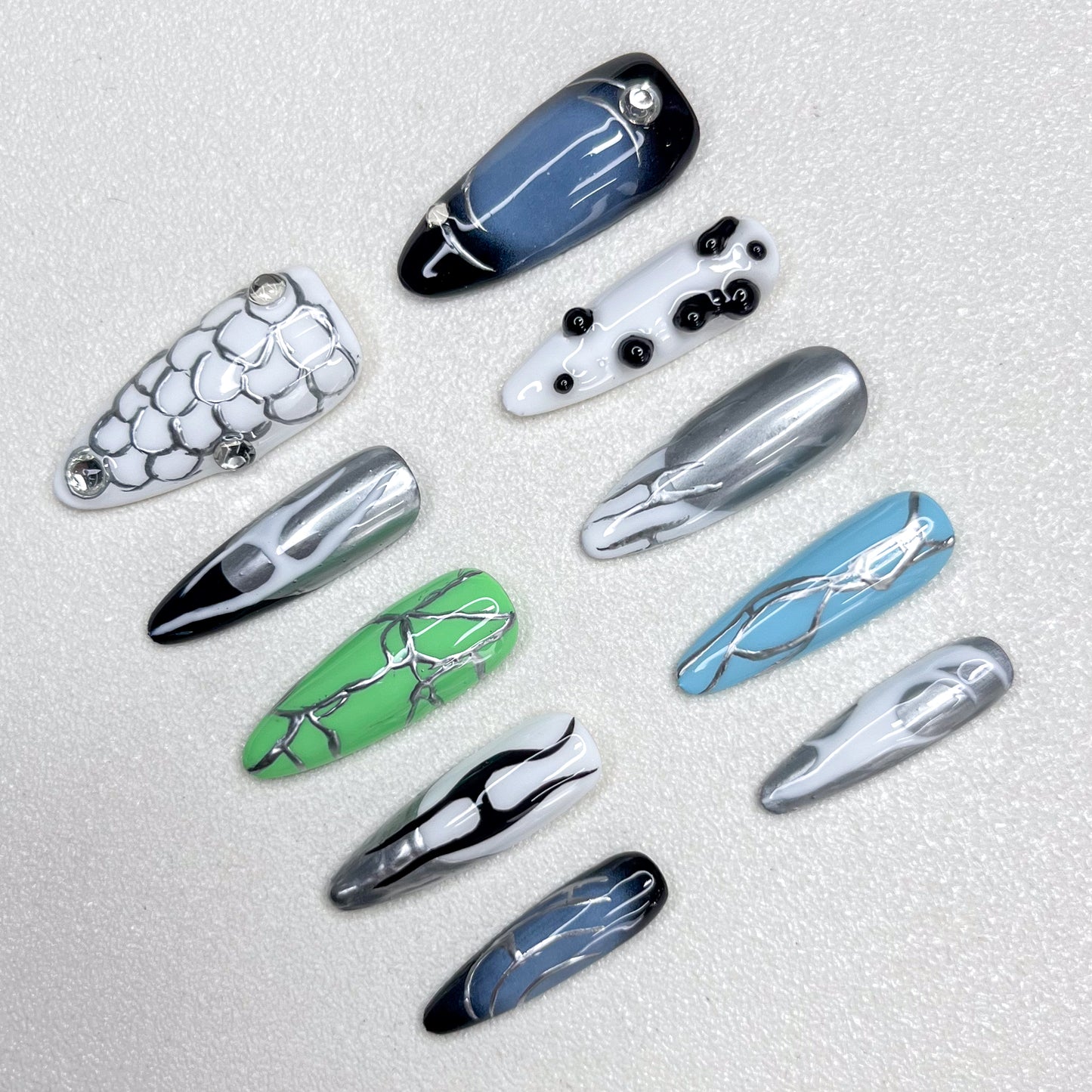Press-on nails inspired by cyberpunk aesthetics, showcasing metallic silver veins across glossy black, green, blue, and white bases, accented with chrome details and geometric patterns for a bold techwear vibe.
