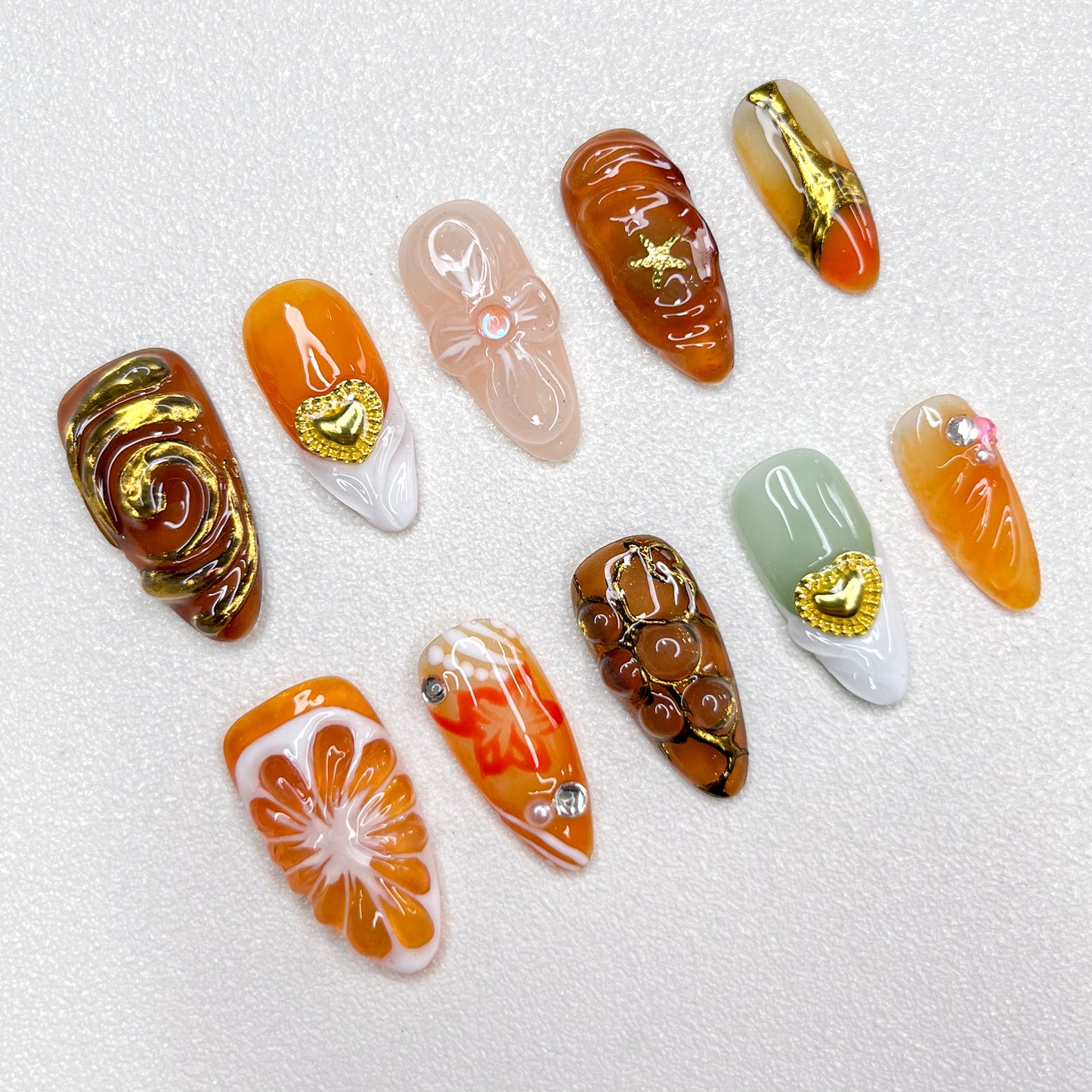 Elegant press-on nails with orange slice accents, gold swirls, heart charms, and warm caramel tones, blending fruity freshness with luxury details in a 3D artistic finish.