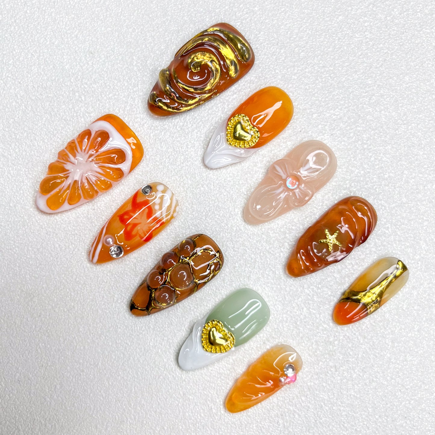 Elegant press-on nails with orange slice accents, gold swirls, heart charms, and warm caramel tones, blending fruity freshness with luxury details in a 3D artistic finish.