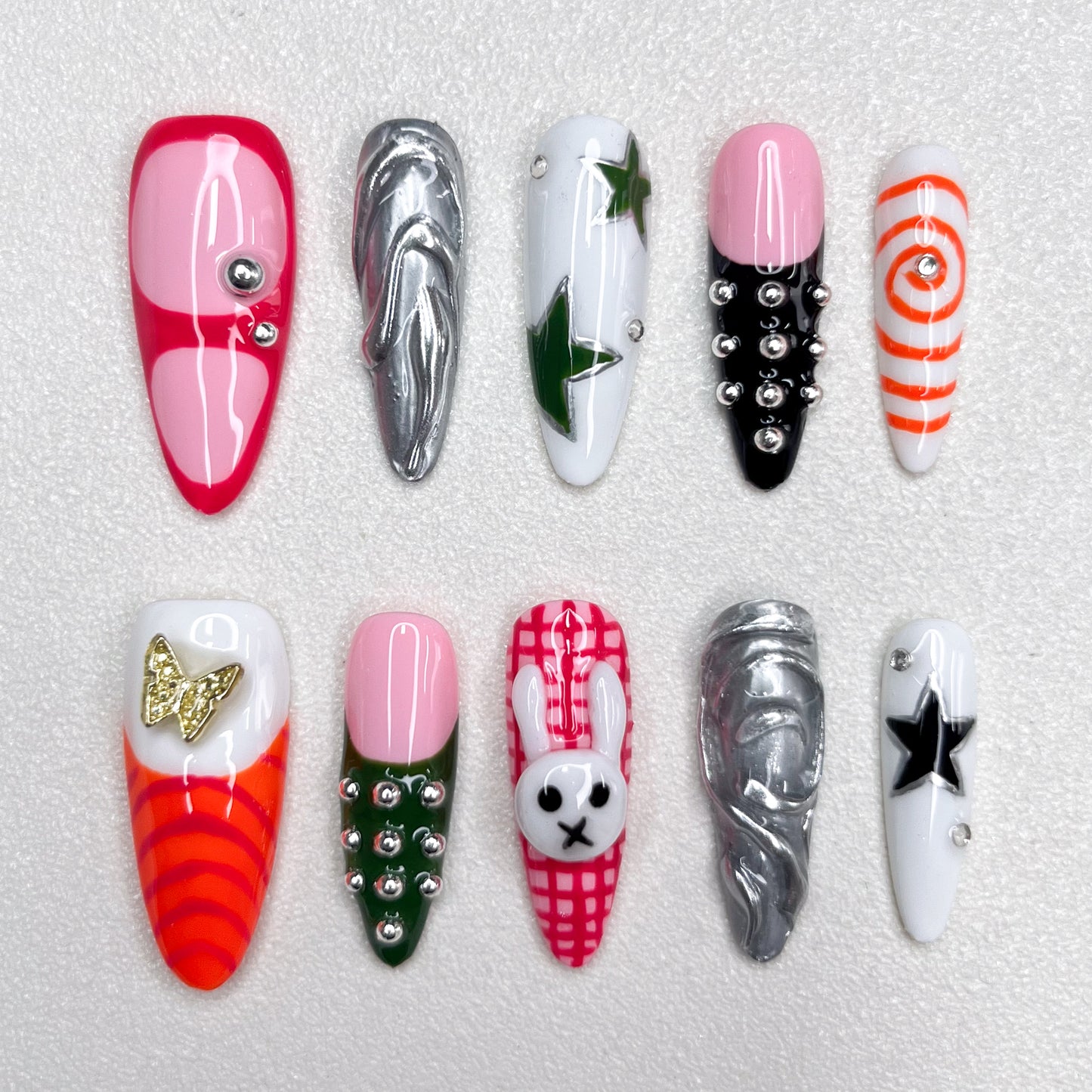 Colorful press-on nails with a mix of stars, spirals, plaid bunny patterns, chrome swirls, and metallic studs—fun, edgy, and fashion-forward Y2K-inspired designs.