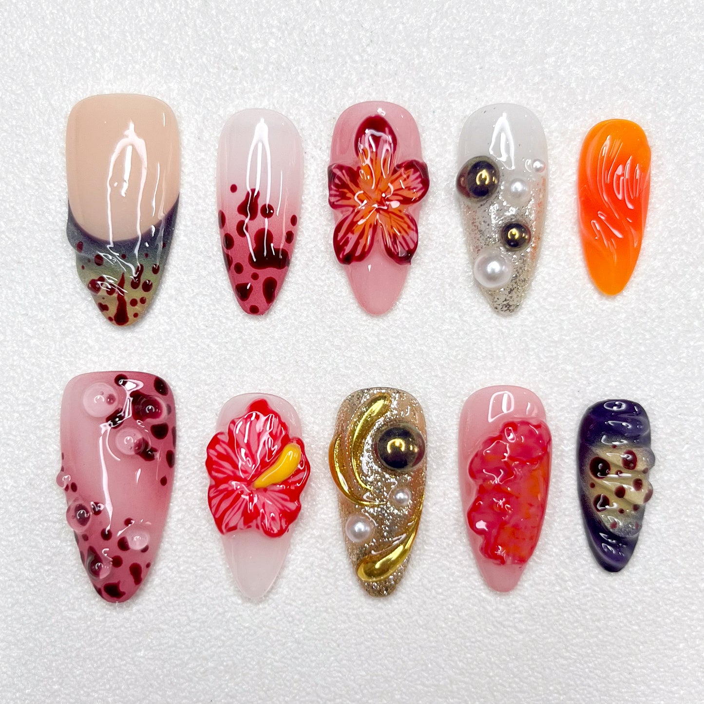 Press-on nails with lava-inspired gradients, red hibiscus flowers, metallic gold swirls, and bubble textures. Includes pearl embellishments and fire-orange, deep wine, and ash grey accents, evoking volcanic eruption and floral bloom.