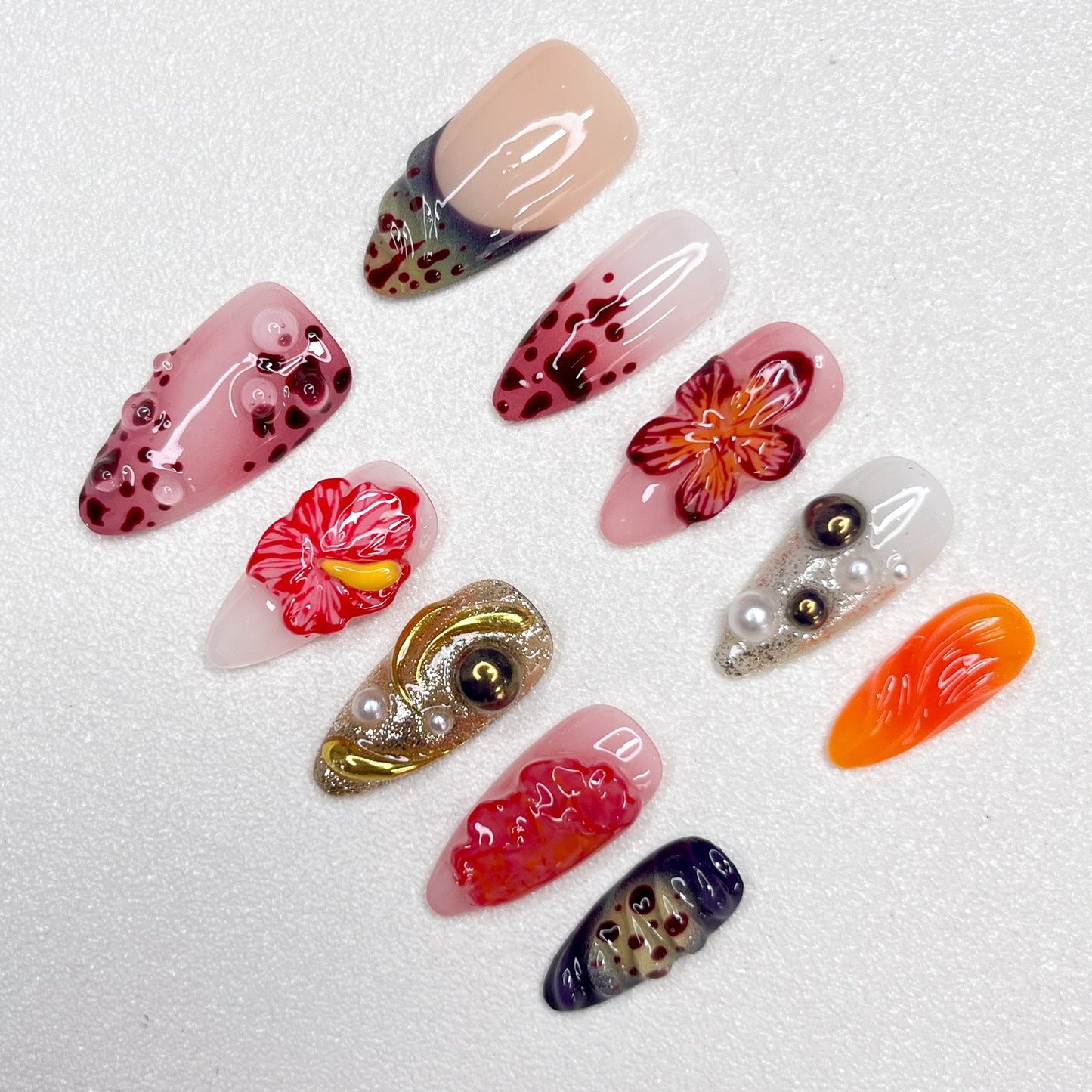 Press-on nails with lava-inspired gradients, red hibiscus flowers, metallic gold swirls, and bubble textures. Includes pearl embellishments and fire-orange, deep wine, and ash grey accents, evoking volcanic eruption and floral bloom.