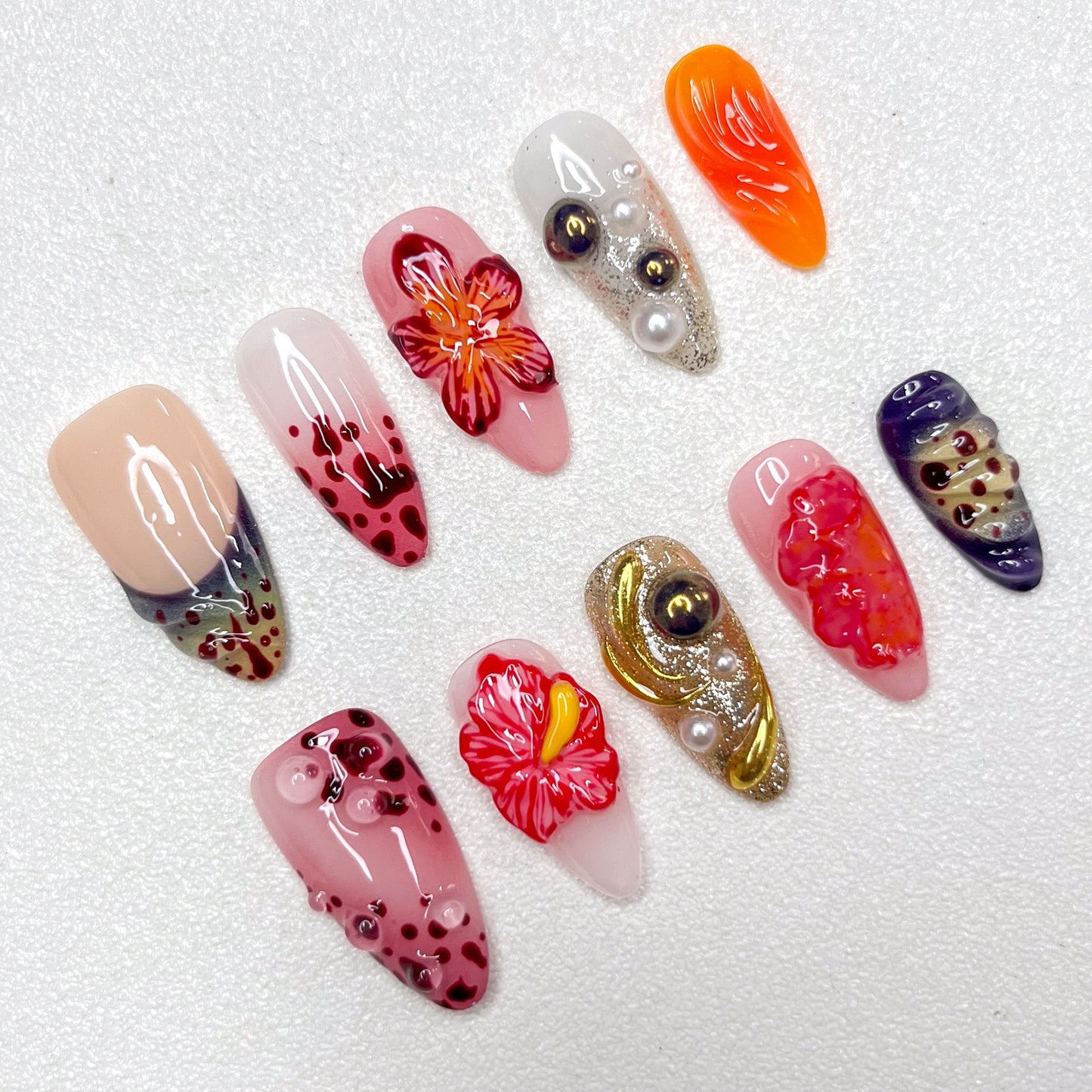 Press-on nails with lava-inspired gradients, red hibiscus flowers, metallic gold swirls, and bubble textures. Includes pearl embellishments and fire-orange, deep wine, and ash grey accents, evoking volcanic eruption and floral bloom.