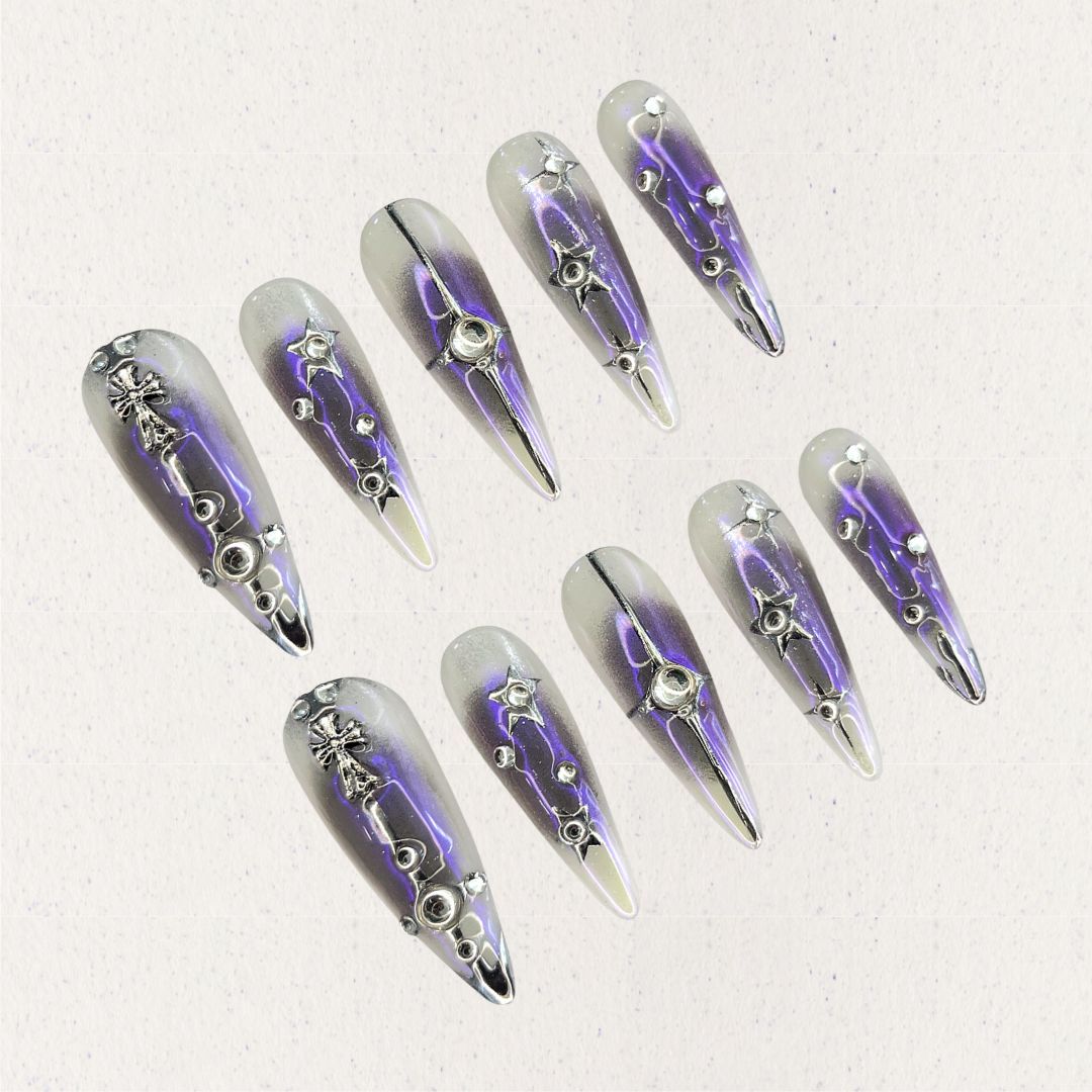 Celestial Radiance presents gradient silver and purple press-on nails with metallic embellishments, including stars and crosses, evoking a celestial and sophisticated style.