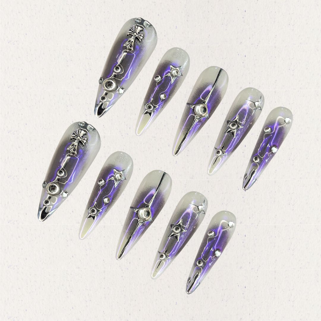 Celestial Radiance presents gradient silver and purple press-on nails with metallic embellishments, including stars and crosses, evoking a celestial and sophisticated style.