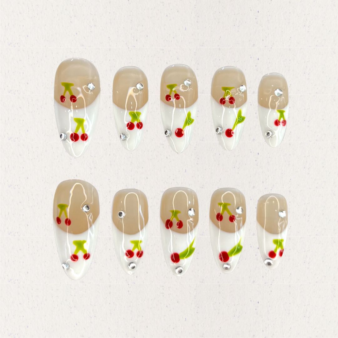 Cherry Charm press-on nails featuring delicate cherry designs on a glossy nude and white base, enhanced with sparkling rhinestones for a cheerful and vibrant aesthetic.