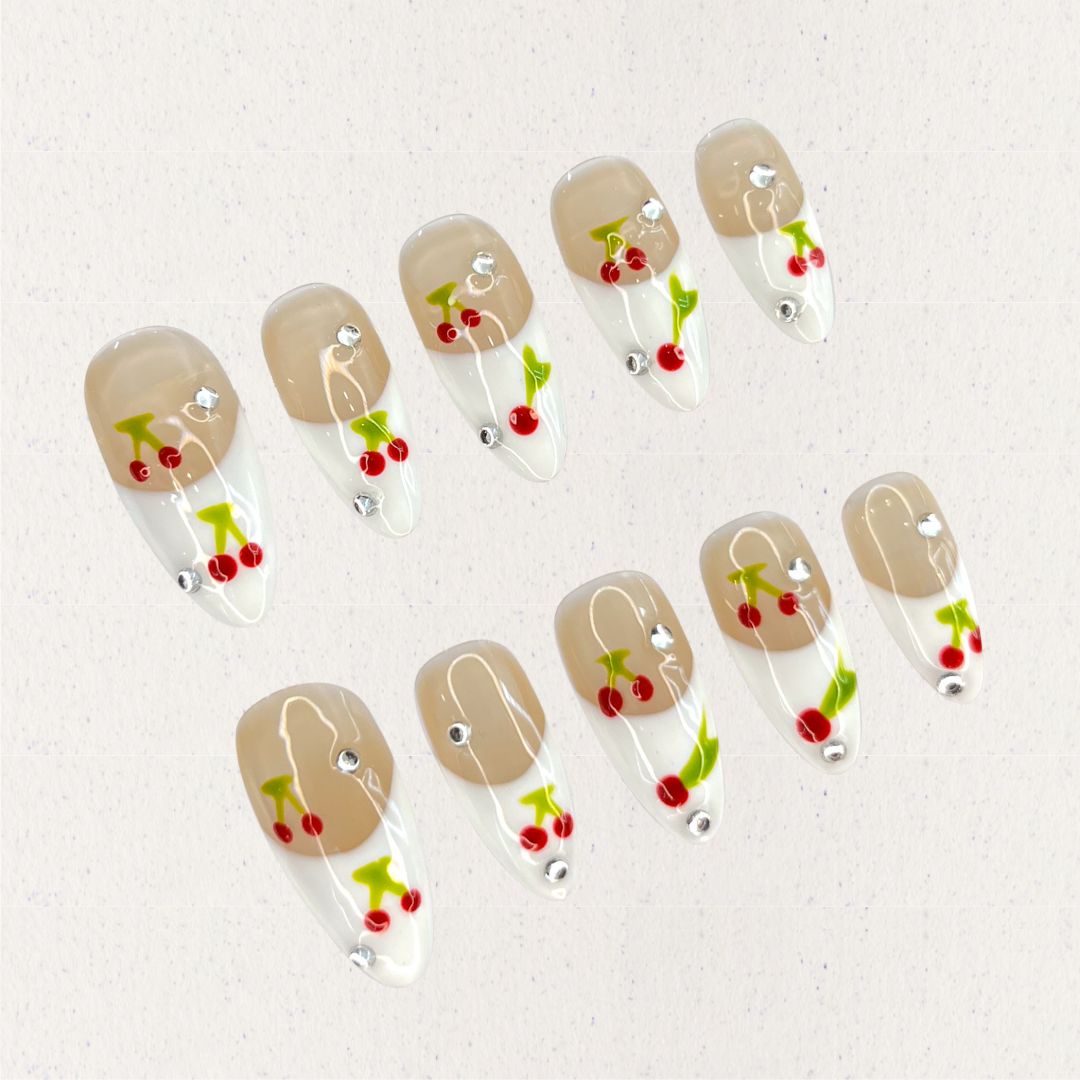 Cherry Charm press-on nails featuring delicate cherry designs on a glossy nude and white base, enhanced with sparkling rhinestones for a cheerful and vibrant aesthetic.