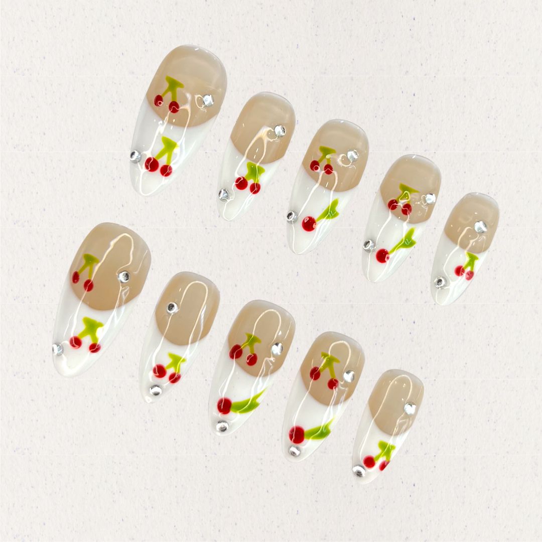 Cherry Charm press-on nails featuring delicate cherry designs on a glossy nude and white base, enhanced with sparkling rhinestones for a cheerful and vibrant aesthetic.