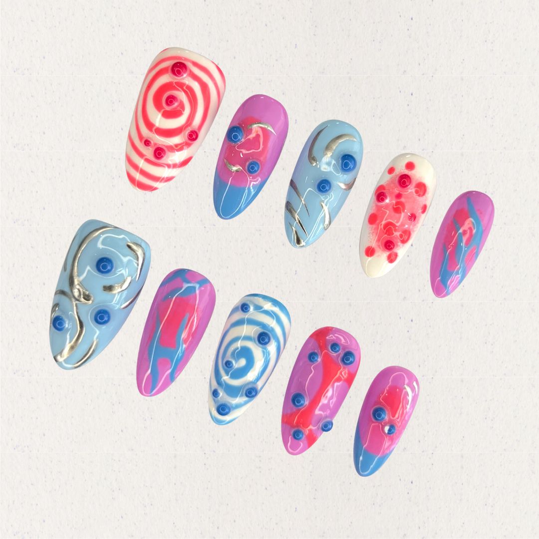 Cosmic Swirls nail set features bold swirls of pink, blue, and white, accented with metallic details and circular 3D elements. This playful yet elegant design mirrors the energetic flow of cosmic waves and nebulae.