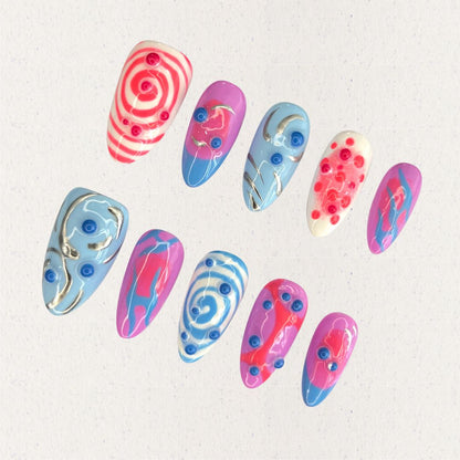 Cosmic Swirls nail set features bold swirls of pink, blue, and white, accented with metallic details and circular 3D elements. This playful yet elegant design mirrors the energetic flow of cosmic waves and nebulae.