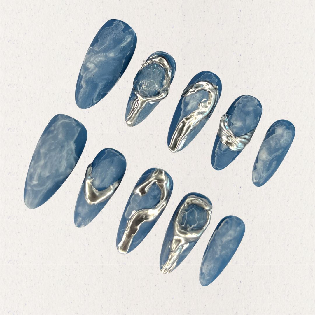 This press-on nail set, titled "Metallic Tide," showcases a deep ocean blue base with delicate marbled textures, resembling swirling water patterns. The standout silver metallic accents create fluid, wave-like designs that mimic flowing liquid metal. The overall aesthetic is modern, minimalist, and artistic, blending nature's elegance with a futuristic touch.