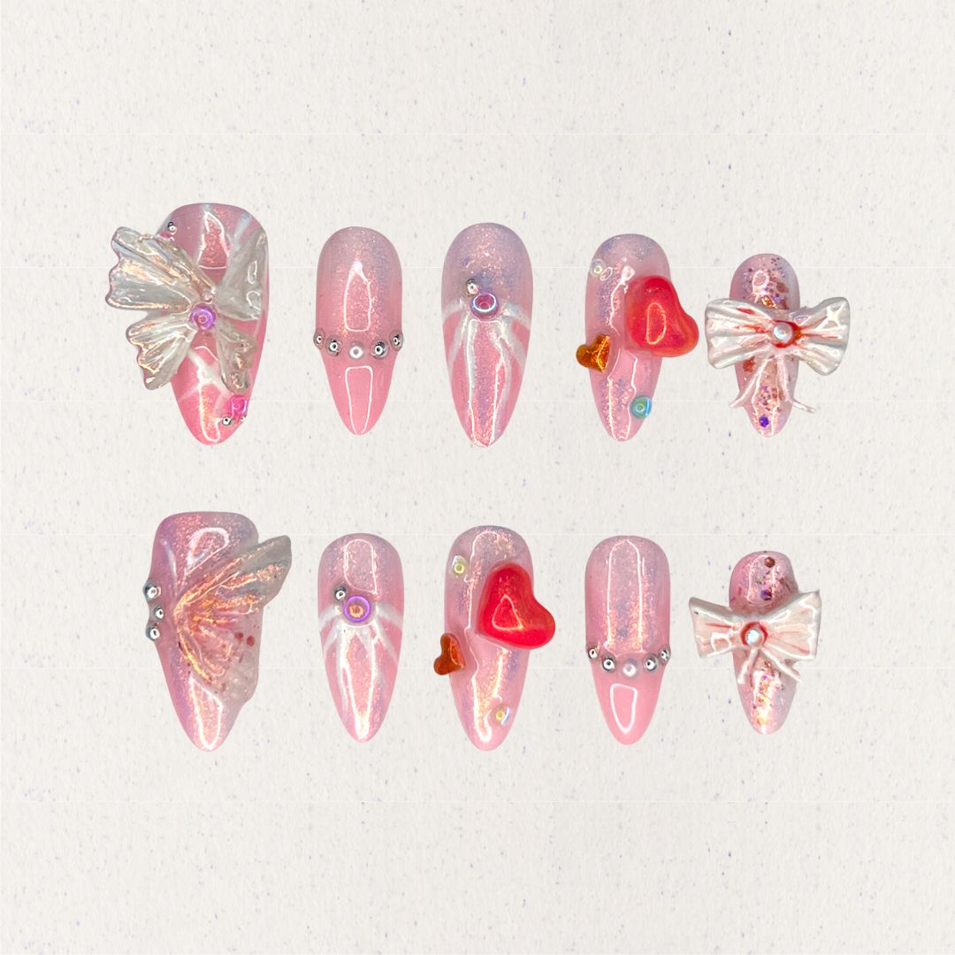 Angelic Grace nail set features shimmering pink hues paired with 3D butterfly wings, intricate ribbons, and vibrant heart accents. This design evokes the elegance of angelic beings, capturing a sense of ethereal beauty and charm.