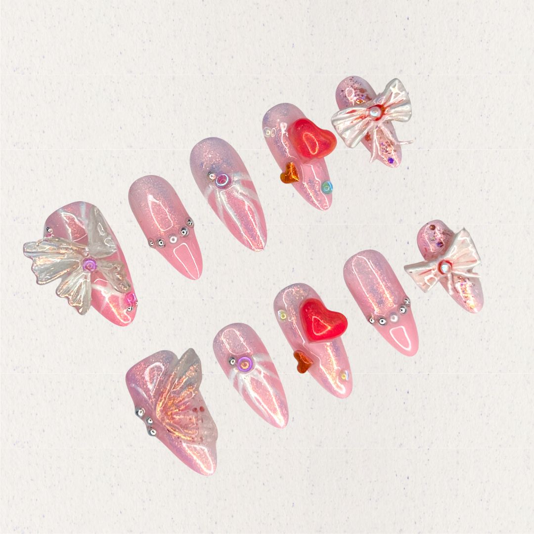 Angelic Grace nail set features shimmering pink hues paired with 3D butterfly wings, intricate ribbons, and vibrant heart accents. This design evokes the elegance of angelic beings, capturing a sense of ethereal beauty and charm.