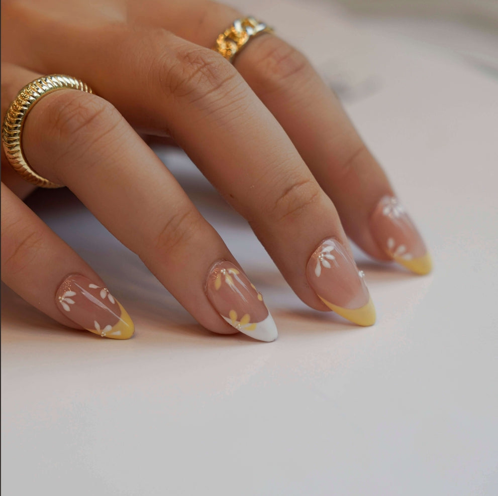 Nude nails with yellow and white French tips, enhanced by delicate daisy-inspired floral accents. Each flower alternates between yellow and white petals with a shimmering diamond at the center, evoking the tranquil beauty of a sunlit meadow.