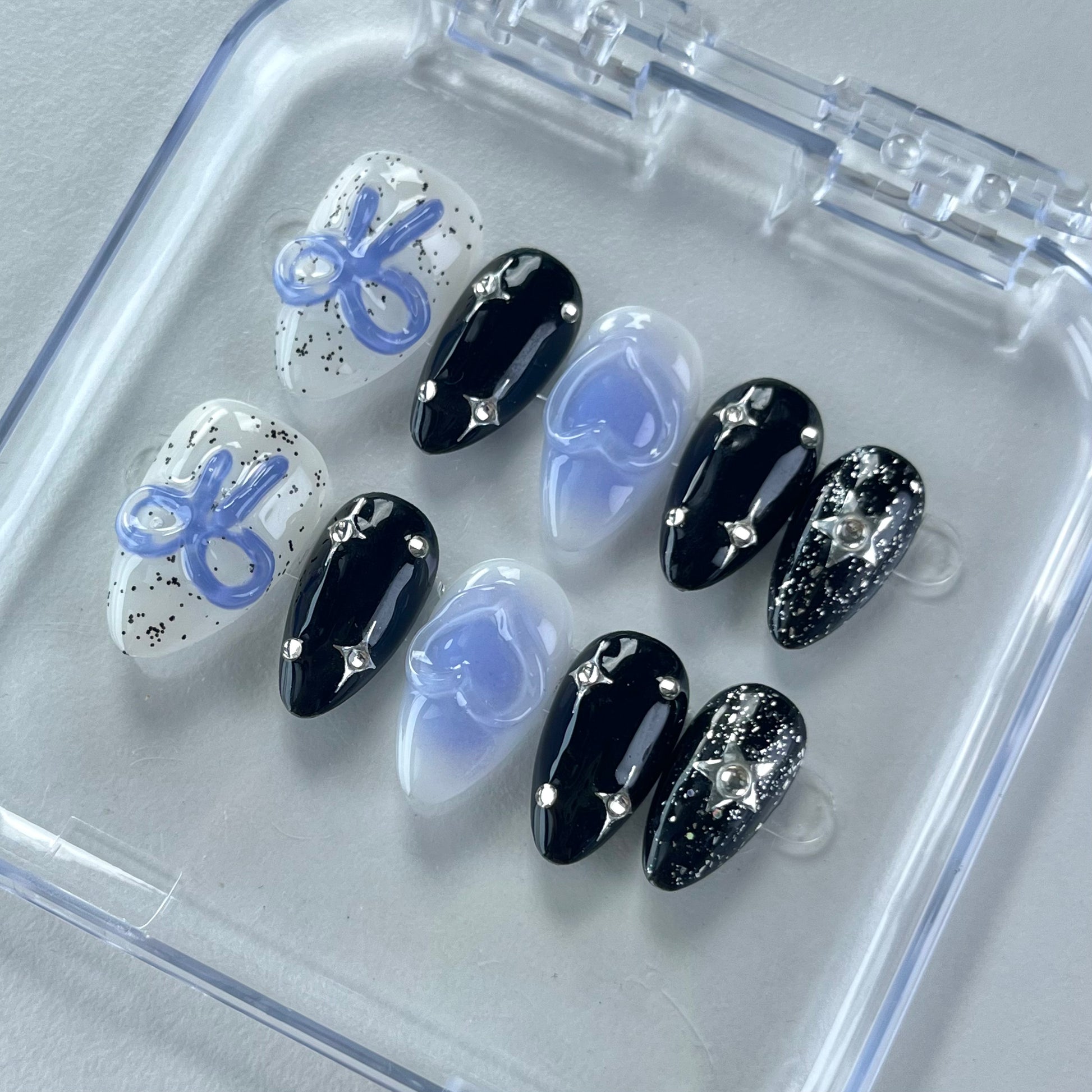 A cosmic-themed press-on nail set with a combination of black and pastel blue nails. The black nails feature glittery silver stars and rhinestones, while the blue nails include elegant bow designs. Perfect for a celestial and dreamy style.