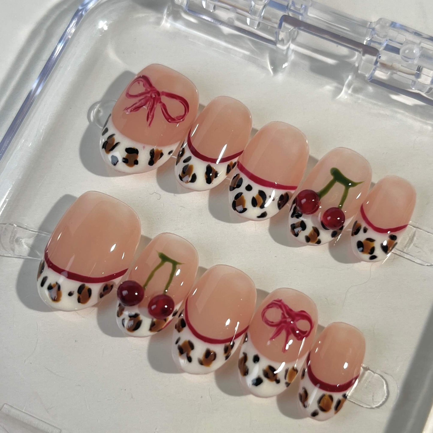 A set of press-on nails with a nude pink base and intricate details. The designs include leopard print French tips, red cherry accents with green stems, and delicate red bows. This artistic nail set blends wild animal motifs with soft, playful touches for a unique fashion statement.