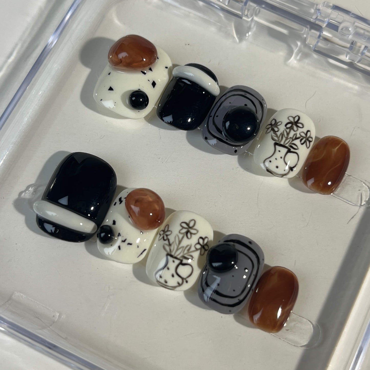 A set of press - on nails with a sophisticated mix of black, white, and brown colors, featuring elegant floral and geometric designs, neatly arranged in a transparent case. The nails have an artistic and stylish finish, ideal for both casual and formal events.