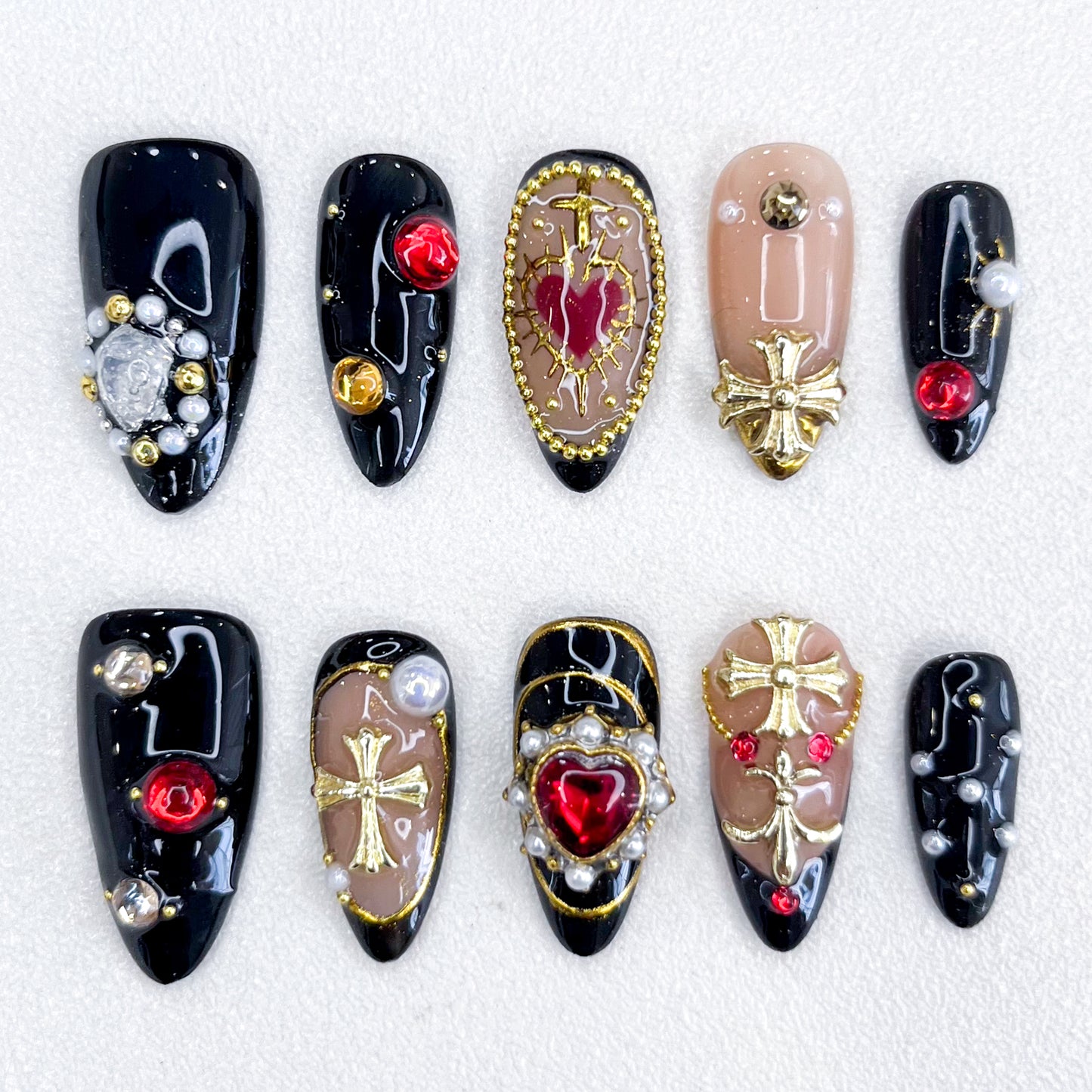 A set of bold and luxurious press-on nails featuring black and nude base tones, embellished with gold cross designs, red heart and round gemstones, pearls, and intricate metallic accents. The design exudes gothic elegance and a regal aesthetic.