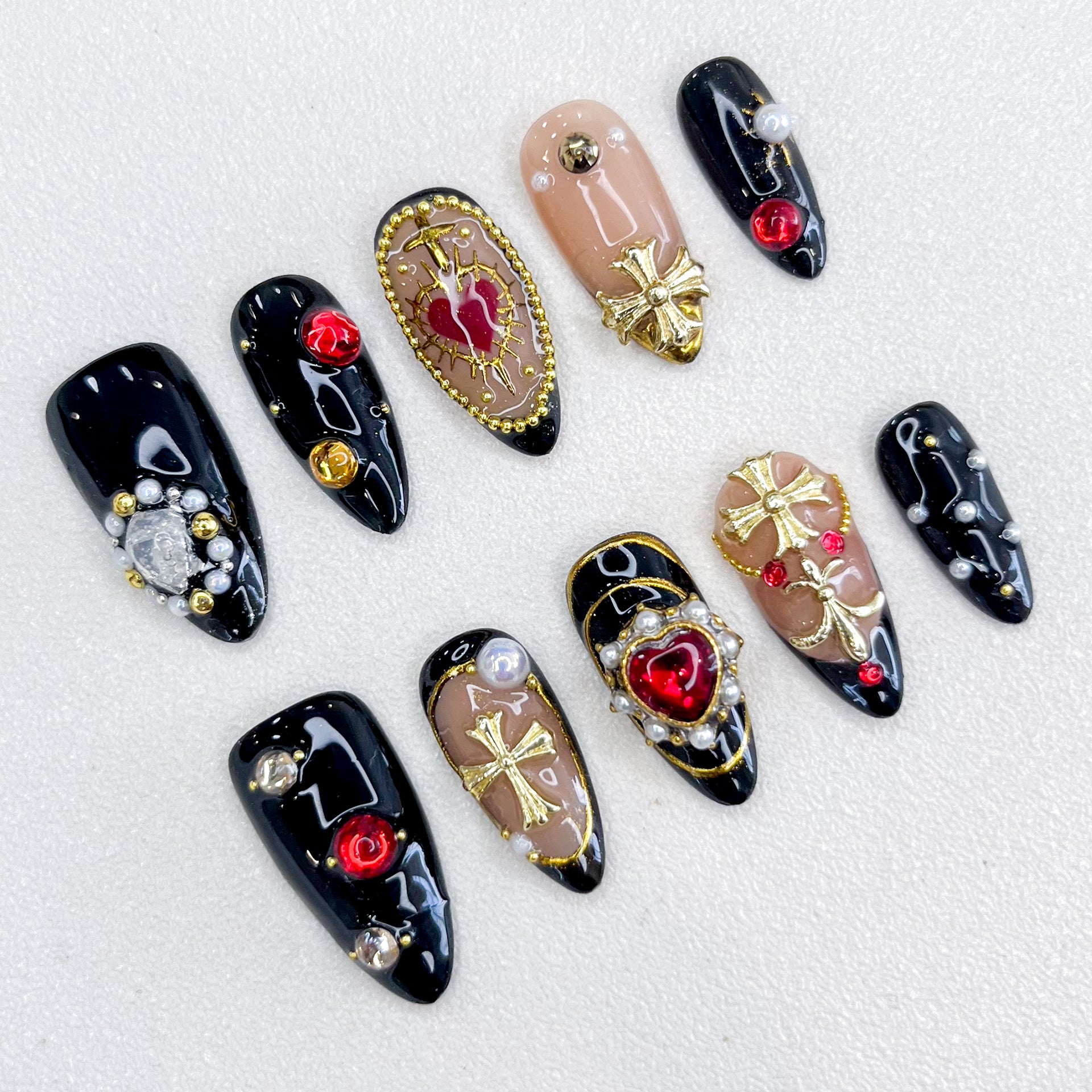 A set of bold and luxurious press-on nails featuring black and nude base tones, embellished with gold cross designs, red heart and round gemstones, pearls, and intricate metallic accents. The design exudes gothic elegance and a regal aesthetic.