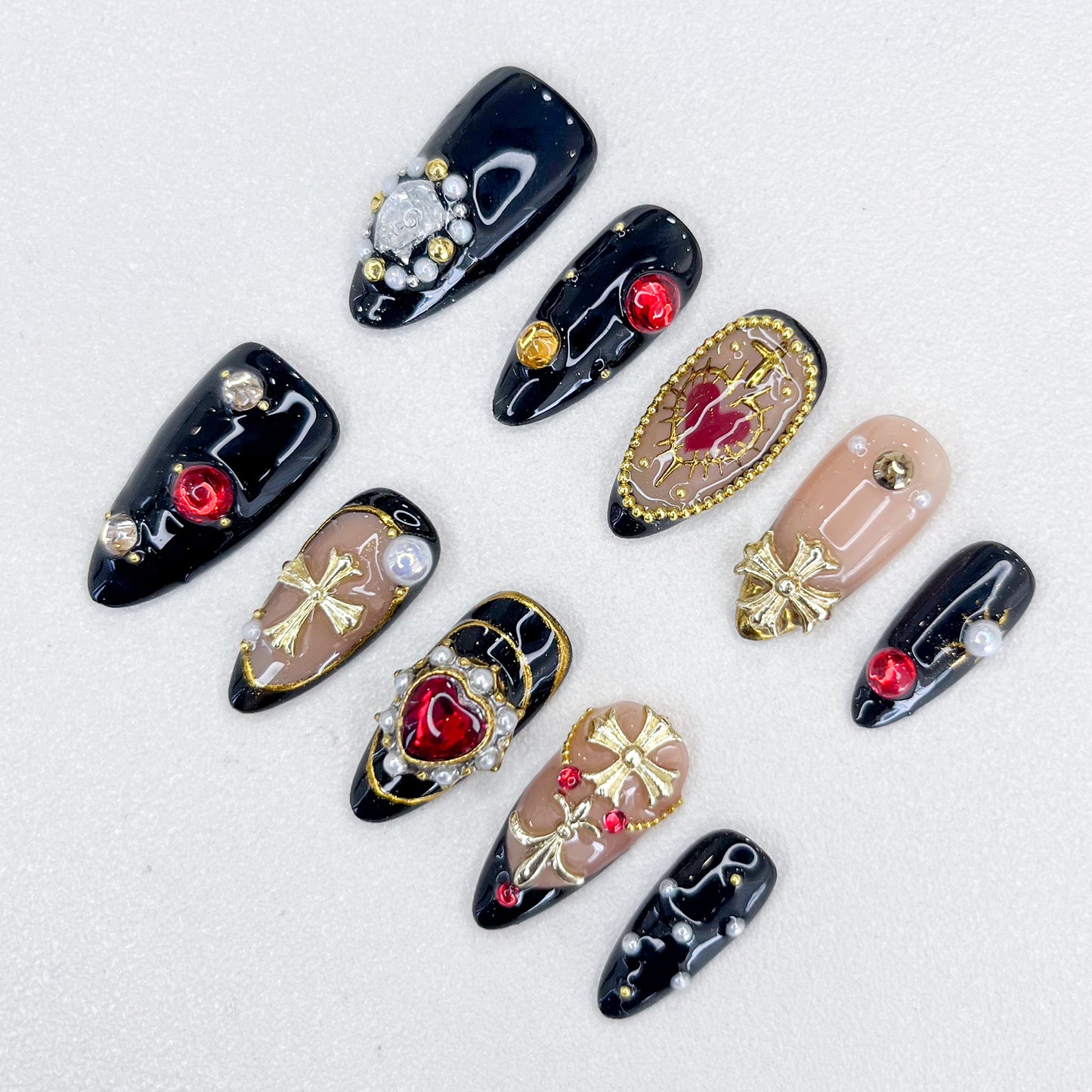 A set of bold and luxurious press-on nails featuring black and nude base tones, embellished with gold cross designs, red heart and round gemstones, pearls, and intricate metallic accents. The design exudes gothic elegance and a regal aesthetic.