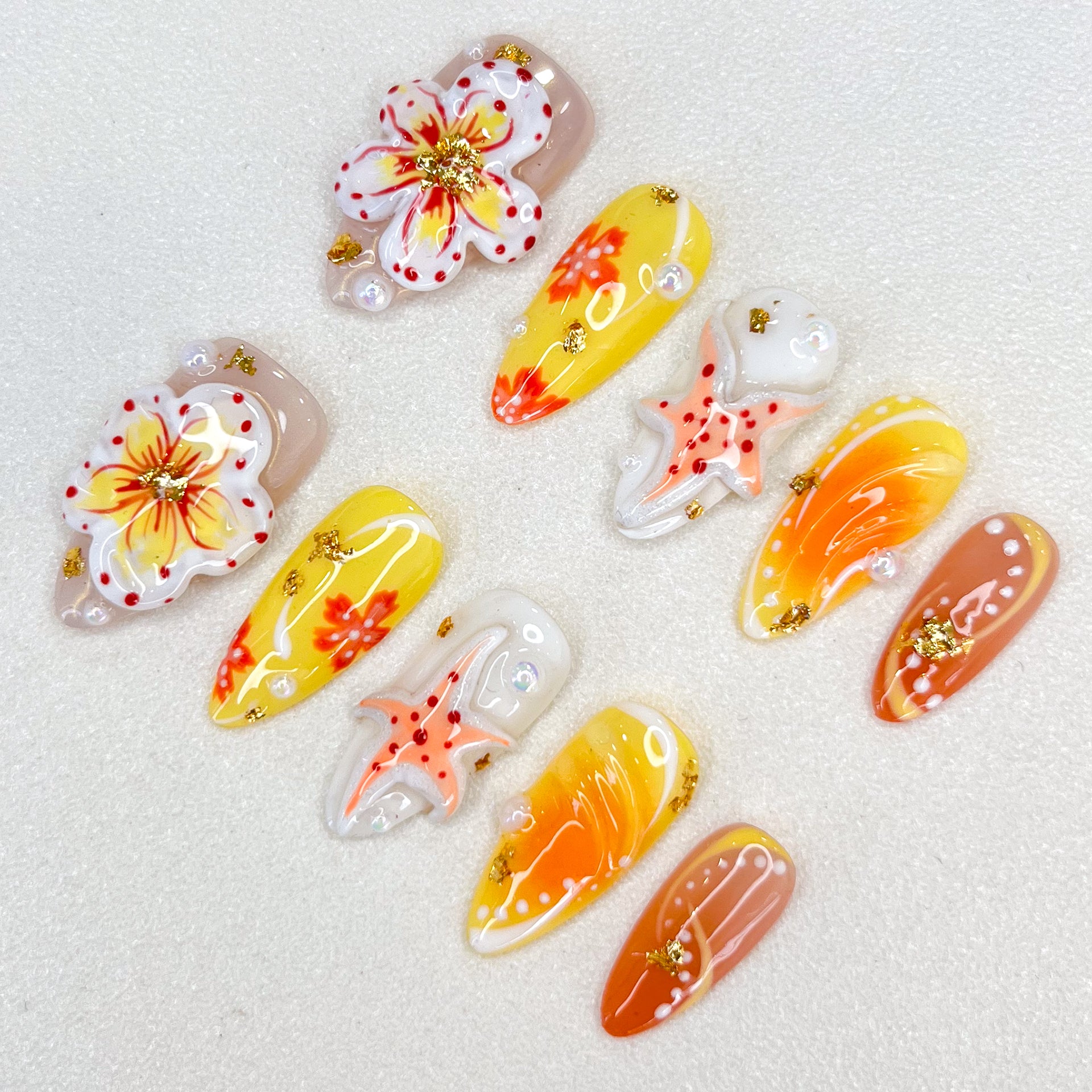 A colorful set of press-on nails with yellow, orange, and nude gradients, adorned with hand-painted flowers, starfish motifs, pearls, and gold foil accents. The design is vibrant and tropical, perfect for summer or beach-themed occasions.