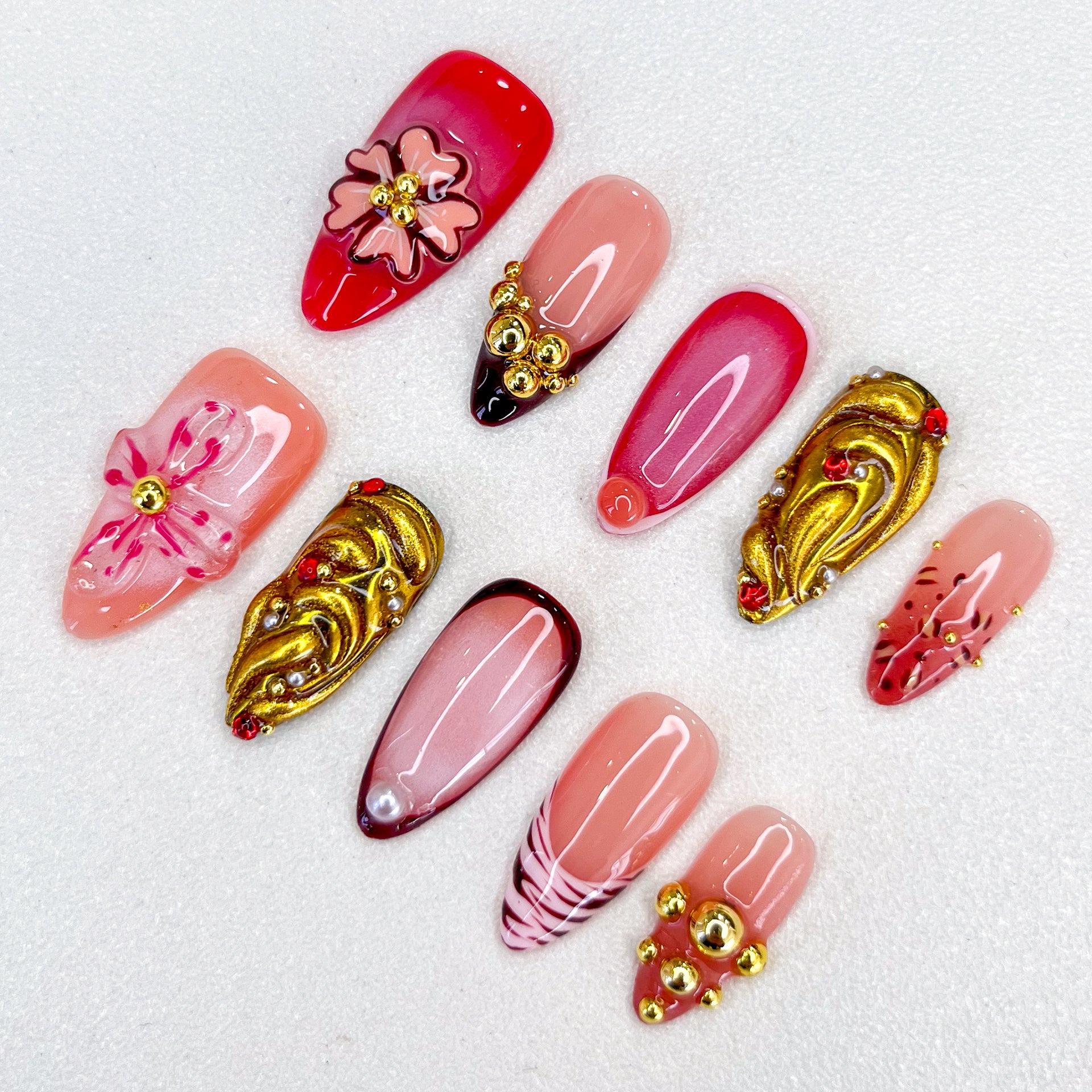 A glamorous set of press-on nails with red, pink, and nude tones, featuring 3D floral embellishments, gold accents, gradient effects, and intricate details. The design is romantic and elegant, perfect for special events or celebrations.