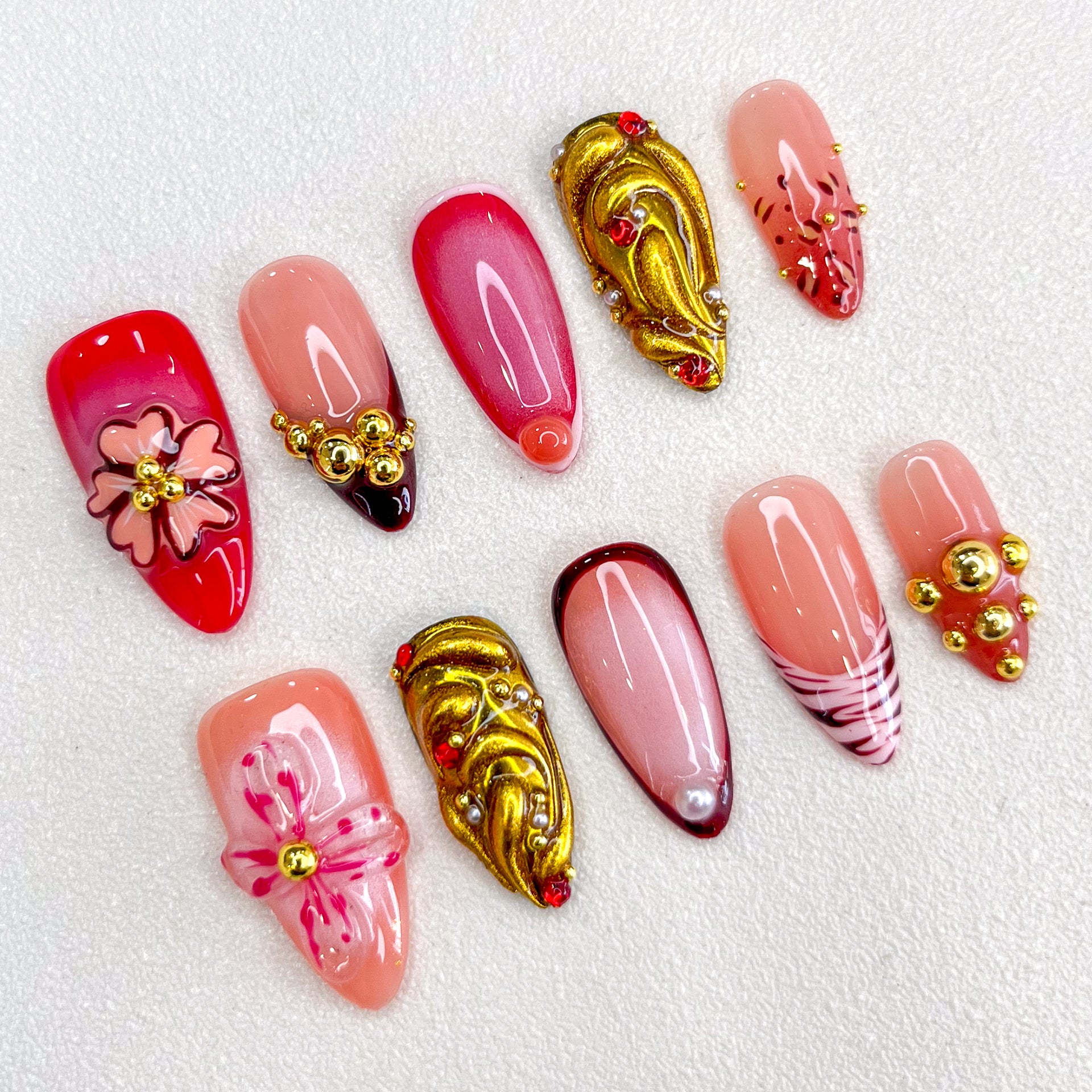 A glamorous set of press-on nails with red, pink, and nude tones, featuring 3D floral embellishments, gold accents, gradient effects, and intricate details. The design is romantic and elegant, perfect for special events or celebrations.