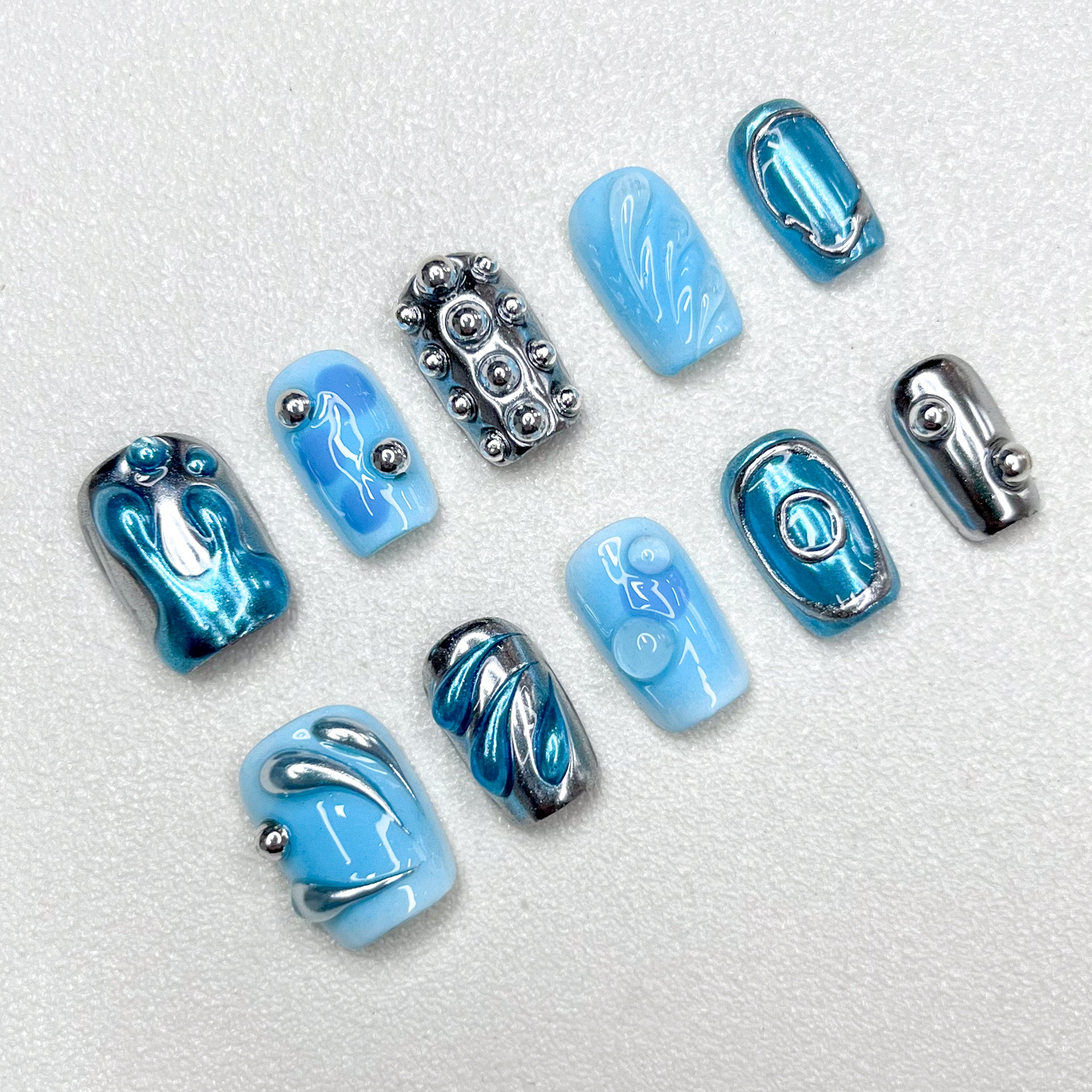 A cyber-inspired press-on nail set in icy blue and metallic chrome tones, featuring 3D orbs, abstract swirls, and smooth reflective patterns. A sleek, sci-fi style manicure with avant-garde vibes.