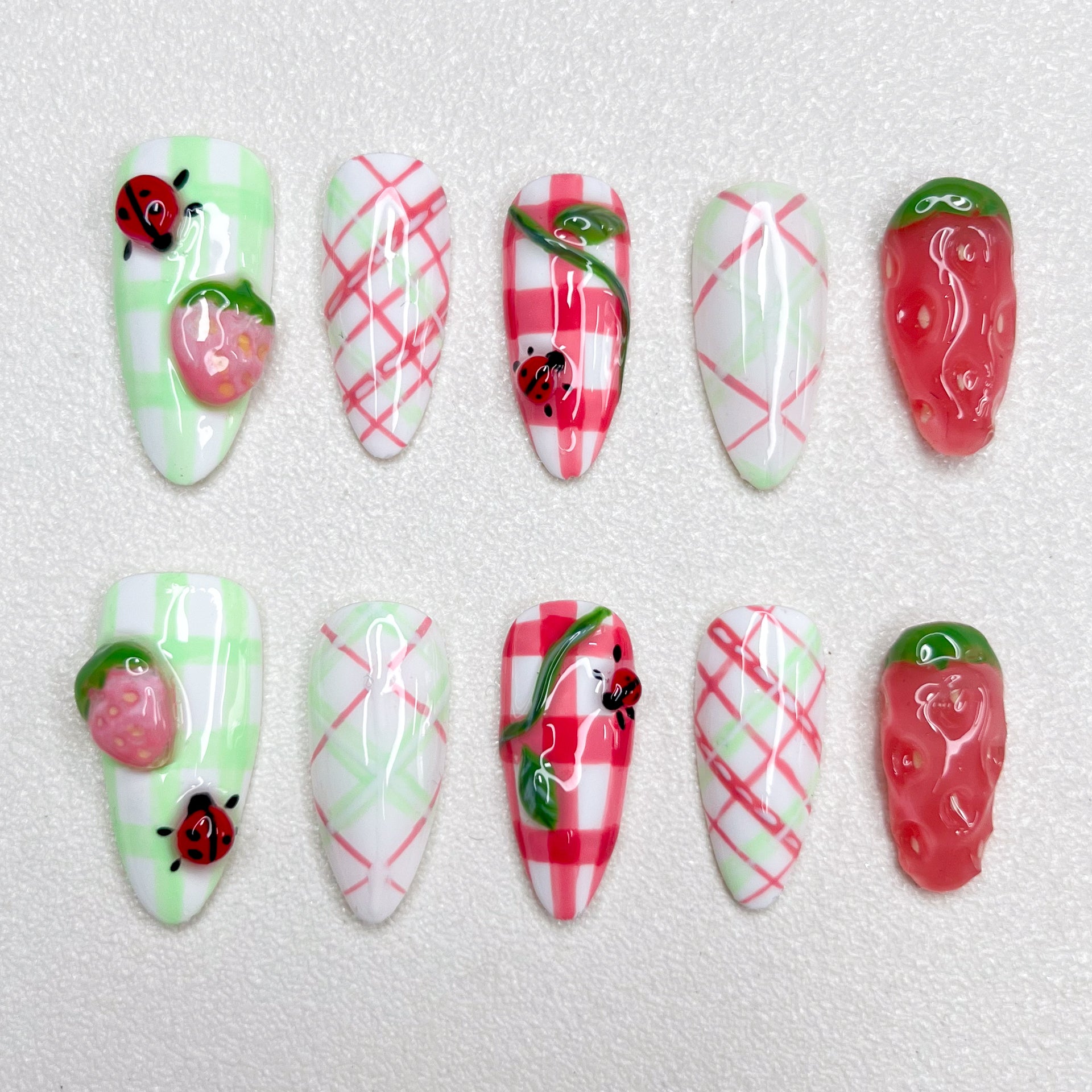 A charming press-on nail set decorated with checkered gingham prints, pink strawberries, and little ladybugs, combining fruity textures and playful summer picnic vibes in green, red, and white hues.