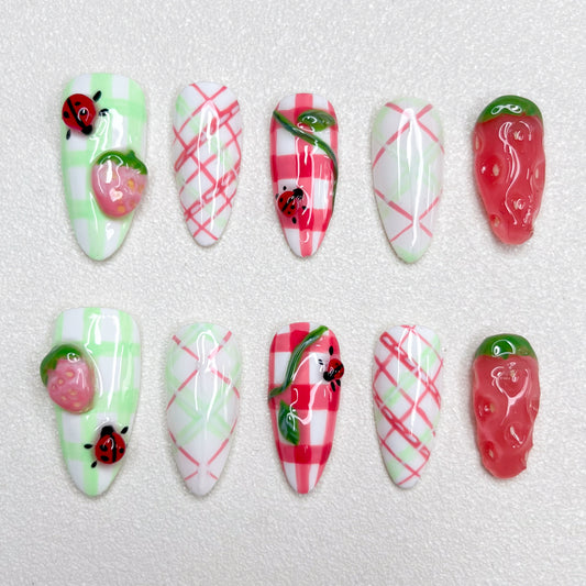 A charming press-on nail set decorated with checkered gingham prints, pink strawberries, and little ladybugs, combining fruity textures and playful summer picnic vibes in green, red, and white hues.
