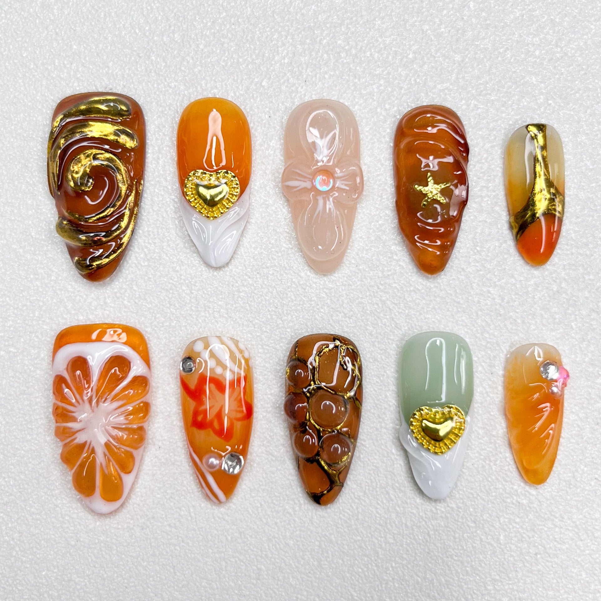 Elegant press-on nails with orange slice accents, gold swirls, heart charms, and warm caramel tones, blending fruity freshness with luxury details in a 3D artistic finish.
