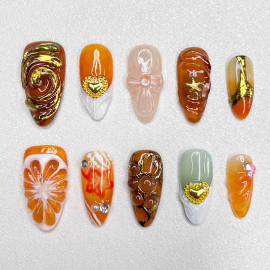 Elegant press-on nails with orange slice accents, gold swirls, heart charms, and warm caramel tones, blending fruity freshness with luxury details in a 3D artistic finish.