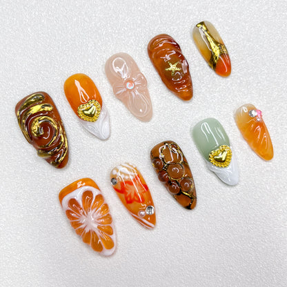 Elegant press-on nails with orange slice accents, gold swirls, heart charms, and warm caramel tones, blending fruity freshness with luxury details in a 3D artistic finish.