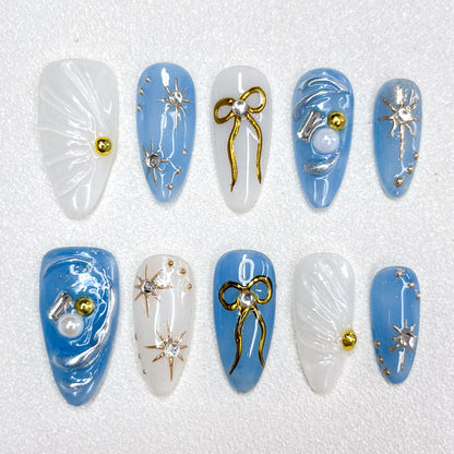 Blue and white celestial-themed press-on nails with 3D golden bows, silver starbursts, pearls, and shimmering accents, evoking a dreamy galaxy-inspired look.