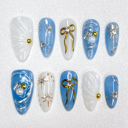 Blue and white celestial-themed press-on nails with 3D golden bows, silver starbursts, pearls, and shimmering accents, evoking a dreamy galaxy-inspired look.