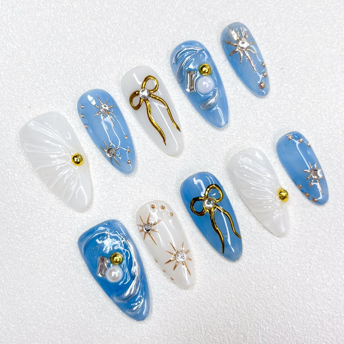 Blue and white celestial-themed press-on nails with 3D golden bows, silver starbursts, pearls, and shimmering accents, evoking a dreamy galaxy-inspired look.
