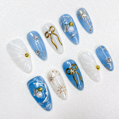 Blue and white celestial-themed press-on nails with 3D golden bows, silver starbursts, pearls, and shimmering accents, evoking a dreamy galaxy-inspired look.
