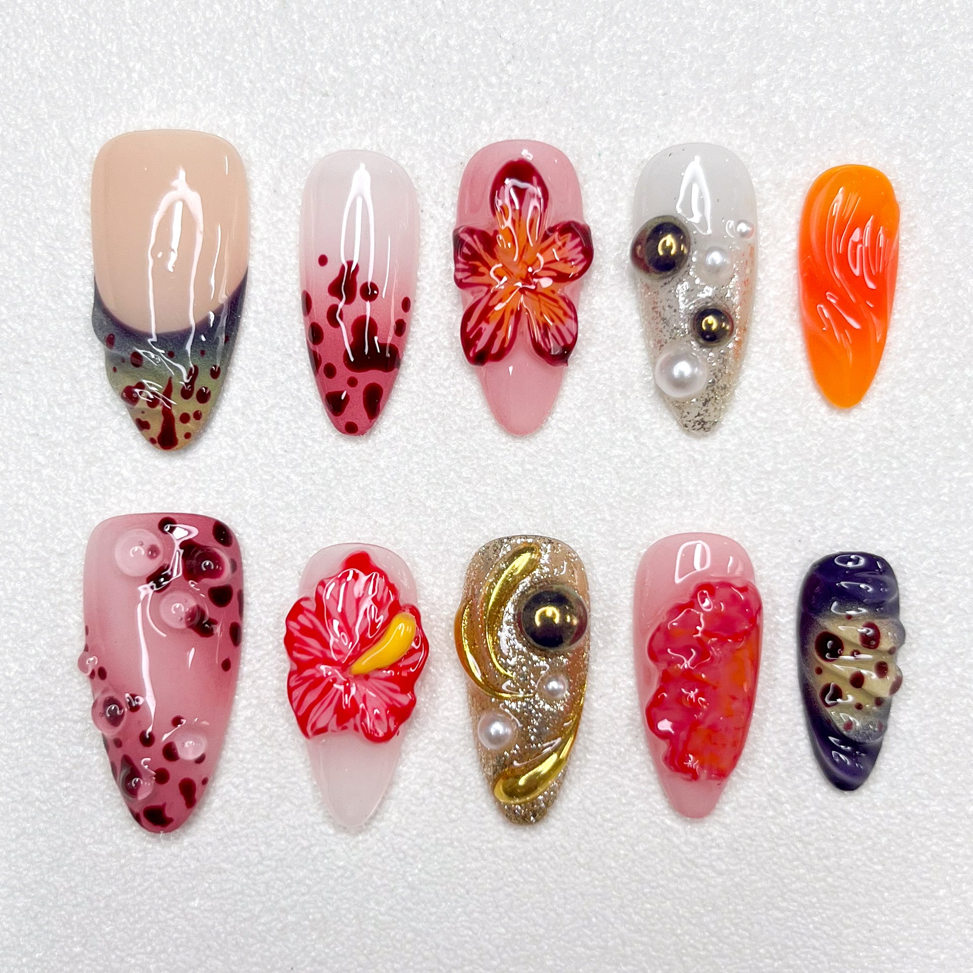 Press-on nails with lava-inspired gradients, red hibiscus flowers, metallic gold swirls, and bubble textures. Includes pearl embellishments and fire-orange, deep wine, and ash grey accents, evoking volcanic eruption and floral bloom.