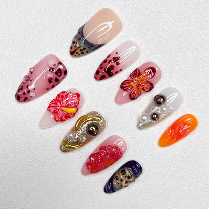 Press-on nails with lava-inspired gradients, red hibiscus flowers, metallic gold swirls, and bubble textures. Includes pearl embellishments and fire-orange, deep wine, and ash grey accents, evoking volcanic eruption and floral bloom.