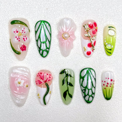 Nature-inspired press-on nails with translucent flowers, gold floral charms, vein-like green outlines, soft pink and white petals, and hand-painted leaves. The design blends garden florals with minimalist elegance in spring tones.