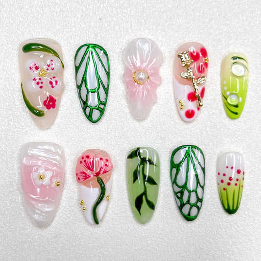 Nature-inspired press-on nails with translucent flowers, gold floral charms, vein-like green outlines, soft pink and white petals, and hand-painted leaves. The design blends garden florals with minimalist elegance in spring tones.
