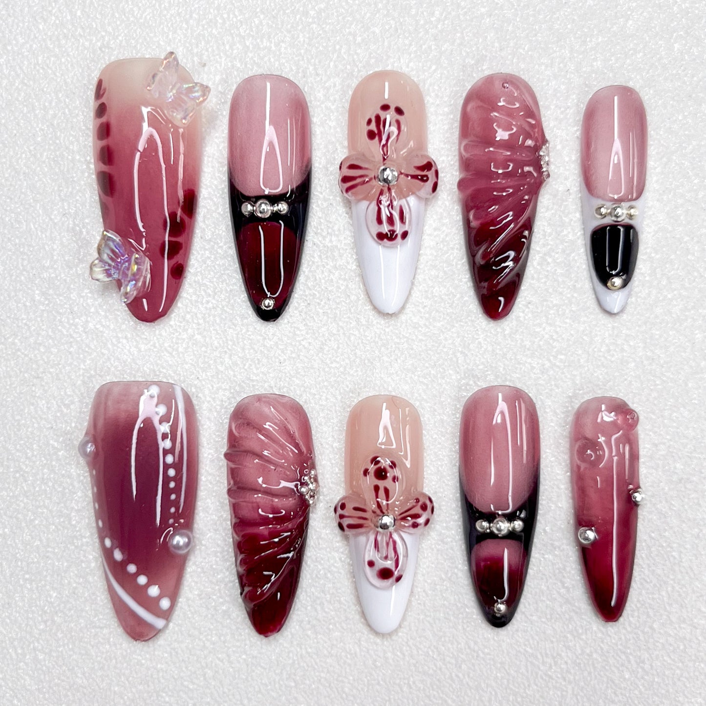 Press-on nails featuring a burgundy-to-black gradient, glossy nude and wine red base tones, 3D butterflies, flower petals, and pearl accents. A luxurious design with gothic elegance and celestial inspiration.