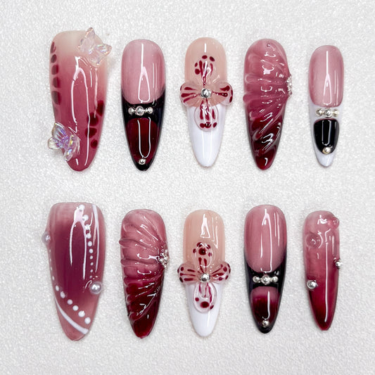 Press-on nails featuring a burgundy-to-black gradient, glossy nude and wine red base tones, 3D butterflies, flower petals, and pearl accents. A luxurious design with gothic elegance and celestial inspiration.