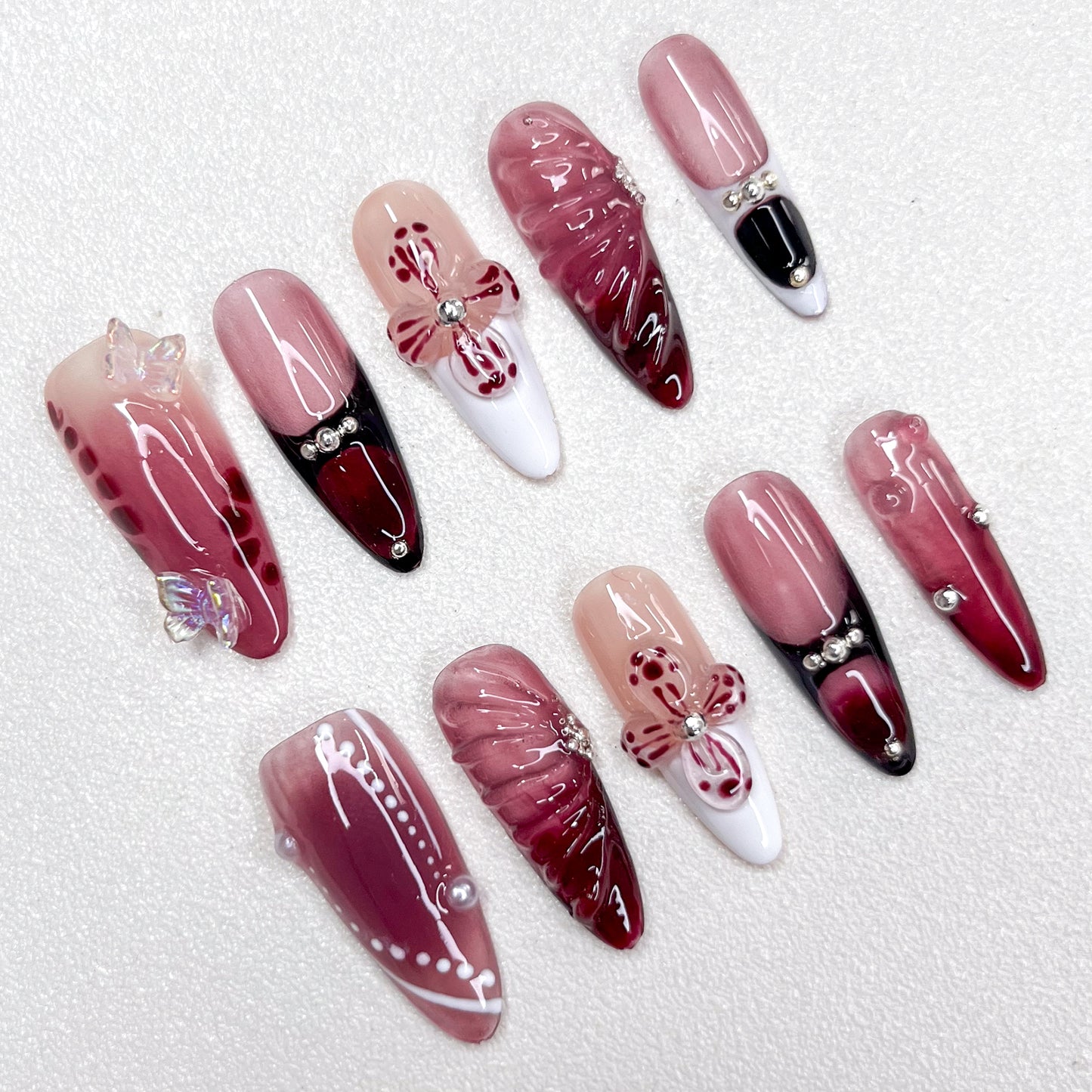 Press-on nails featuring a burgundy-to-black gradient, glossy nude and wine red base tones, 3D butterflies, flower petals, and pearl accents. A luxurious design with gothic elegance and celestial inspiration.